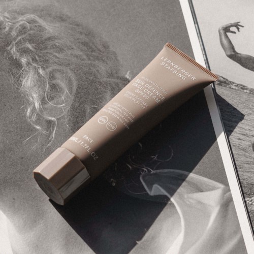 Eine Tube Gesichtspflegecreme liegt auf einer schwarz-weißen Magazinseite neben lockigem Haar und einer künstlerischen Skizze.