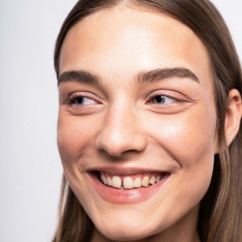 Eine junge Frau mit langem braunen Haar lächelt und schaut nach rechts. Sie hat klare Haut und helle Augen.