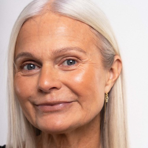 Eine ältere Frau mit langen weißen Haaren, heller Hautfarbe und blauen Augen, lächelt sanft. Sie trägt Ohrringe und hat einen ruhigen Ausdruck.