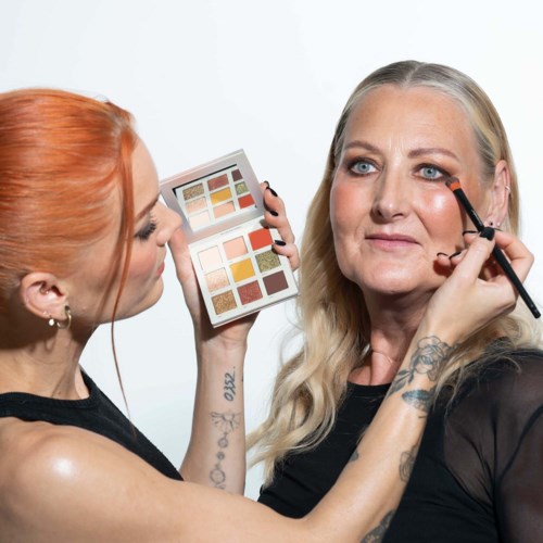 Eine Frau trägt Make-up auf eine andere Frau auf, während sie eine Lidschattenpalette hält.