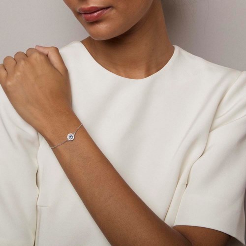 A woman wears a white dress and a delicate bracelet on her wrist, with her arm raised slightly.