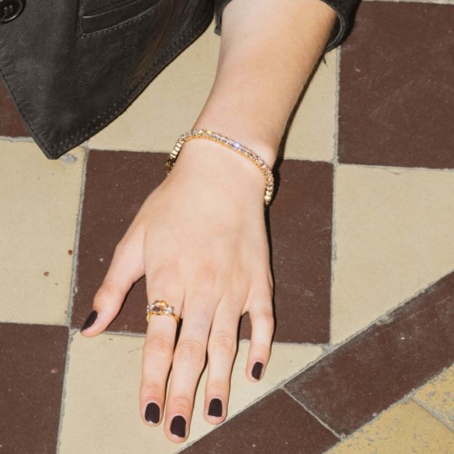 Hand with dark nail polish, gold bracelet, and ring resting on tiled floor.