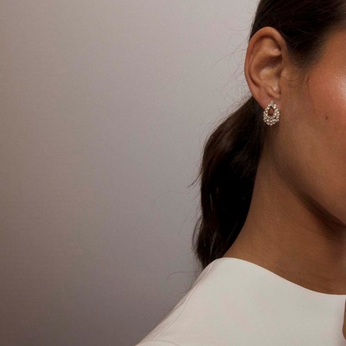 A woman with brown hair in a ponytail, wearing a white top and sparkling earrings.