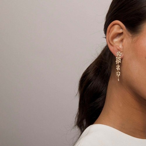 A woman wears a sparkling, long dangling earring and has dark hair tied back. She wears a white top.