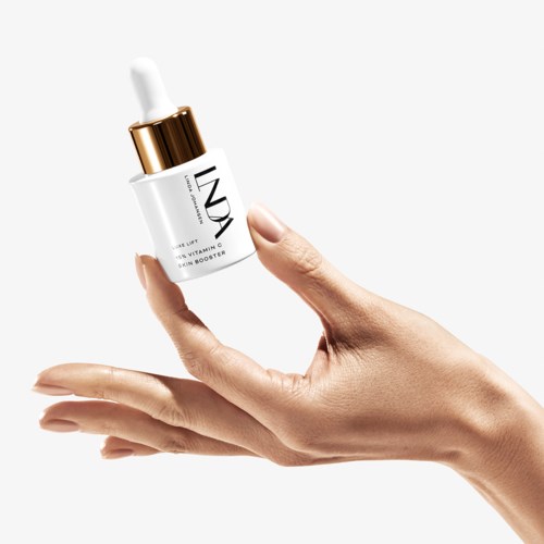 En hånd holder en liten hvit flaske med en pipette, merket 'INDY' og 'Vitamin C Serum Booster'.