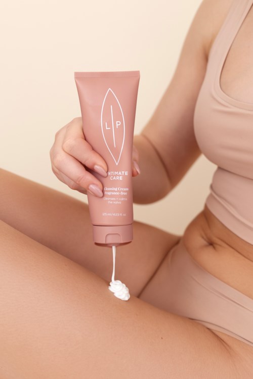 Eine Frau hält eine rosa Tube mit Pflegeprodukt, aus der Creme kommt, sitzt mit hochgelegtem Bein.