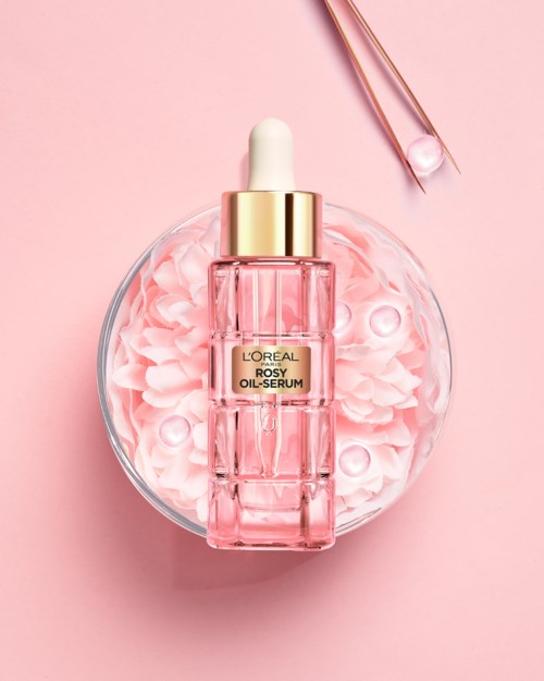 Pink and gold-colored serum bottle on a dish with pink flowers and a pair of pink tweezers.