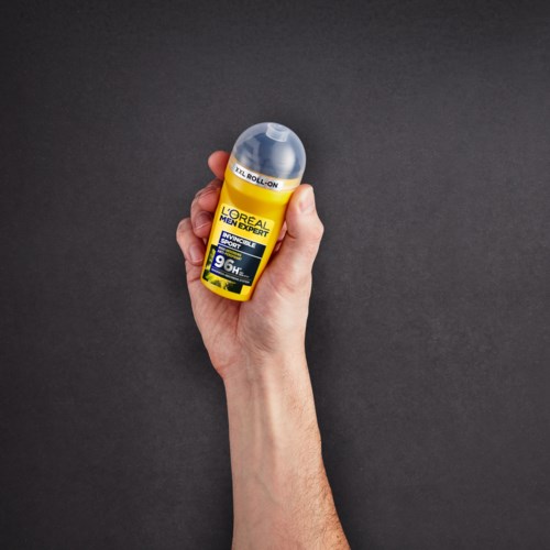 A hand holds a yellow bottle of L'Oreal Men Expert antiperspirant with a roll-on ball, against a dark background.