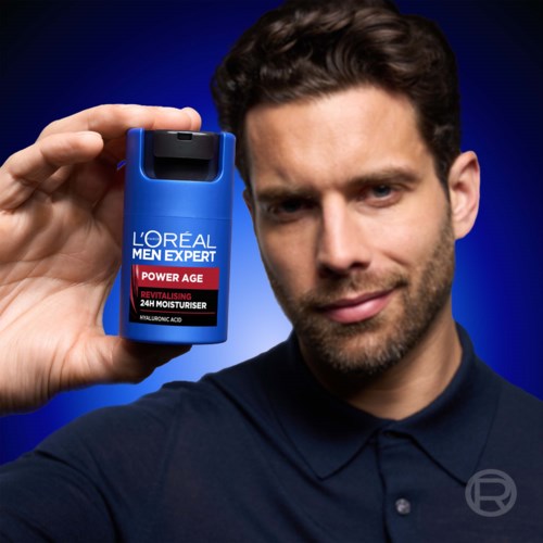 En mann holder en blå flaske Loreal Men Expert Power Age fuktighetskrem nær ansiktet mot en mørkeblå bakgrunn.