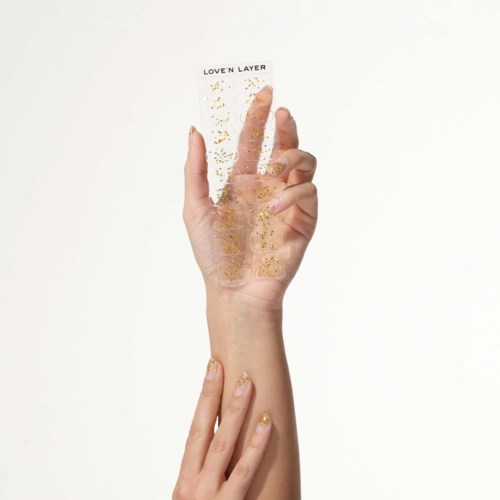 A hand with decorated nails holds a clear sheet titled "Love'n Layer" with gold glitter on a white background.