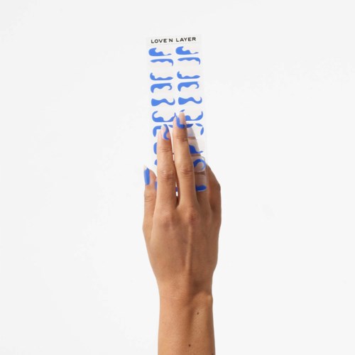 A hand with blue painted nails peels off blue stickers labeled 'LOVEN LAYER' on a white sheet.