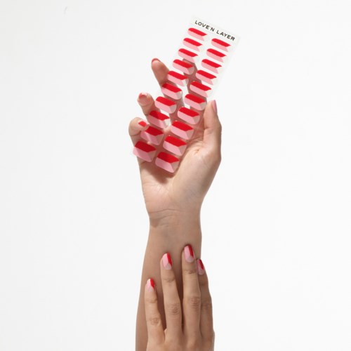 A hand holds a strip of red and pink nails, labeled "LOVE N LAYER," in front of a white background.