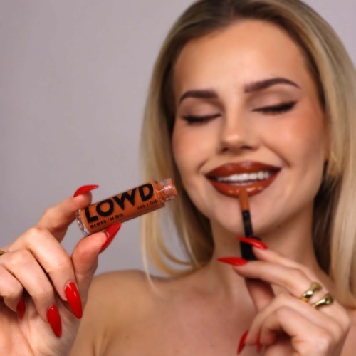 Een vrouw brengt lipstick aan en houdt een lipgloss tube vast. Ze heeft lang blond haar en glimlacht.