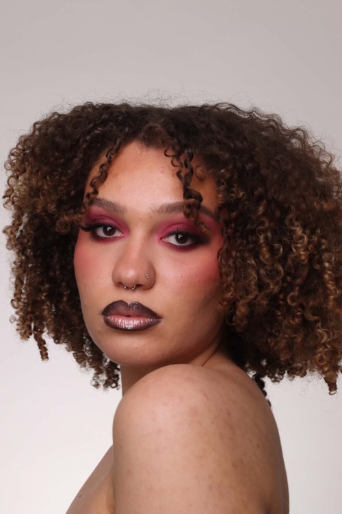 Een vrouw met curly haar, opvallende make-up en een neusring kijkt over haar schouder.