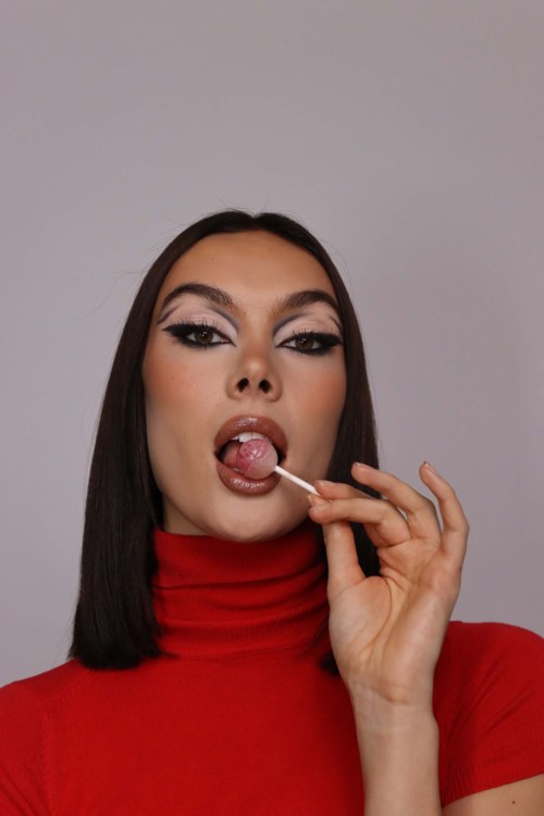 Een vrouw in een rood coltruik likt aan een lolly, met make-up en zwart haar, tegen een eenvoudige achtergrond.