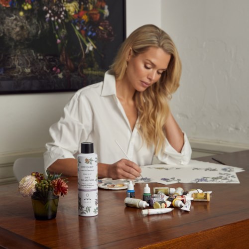 Eine Frau mit langen blonden Haaren malt auf Papier mit verschiedenen Farben. Sie sitzt an einem Holztablett mit Kunstmaterialien.