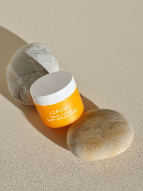 Een kleine gele en witte pot glow crème tussen twee stenen, werpt schaduwen op een beige oppervlak.
