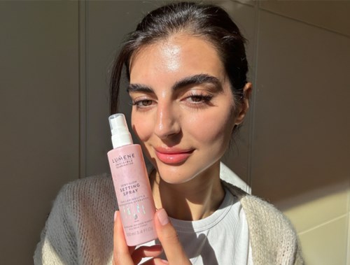 Eine Frau hält eine rosa Flasche Fixierspray, lächelt mit Sonnenlicht im Gesicht.