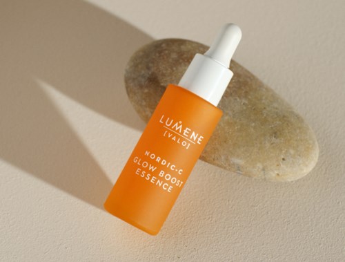 En orange flaska med vit dropperlock märkt 'Lumene IVALO NORDIC-C GLOW BOOST ESSENCE' vilar på en beige yta bredvid en grå sten.