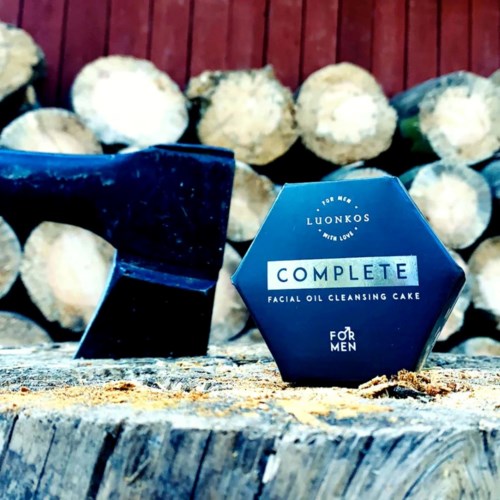 Eine blaue sechsseitige Box mit der Aufschrift "Complete Facial Oil Cleansing Cake" für Männer, mit Holzstämmen im Hintergrund und einem schwarzen Gegenstand in der Nähe.