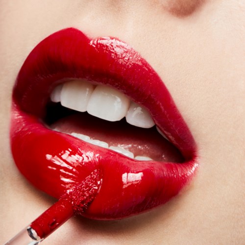Nahaufnahme von Lippen mit rotem Lippenstift, halber offenen Mund, zeigt Zähne, hält roten Lippenstift. Lipgloss ist glänzend.