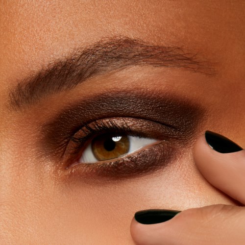 Close-up of a person's eye with dark eyeshadow and well-groomed eyebrows. A hand with black nail polish touches the face.
