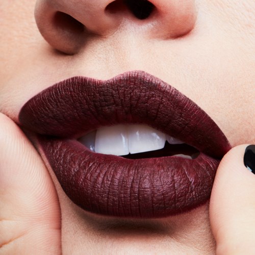 Close-up of lips with dark red lipstick slightly parted, showing teeth, and a finger near the lips.