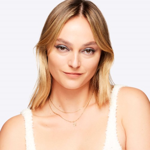 En ung kvinna med blont hår, lätt makeup och ett lugnt leende, iklädd vit topp och lager på lager halsband.