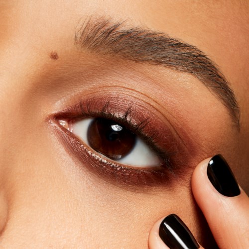 A close-up of a person's eye with makeup, eyebrows, and nails visible, touching the skin below the eye.