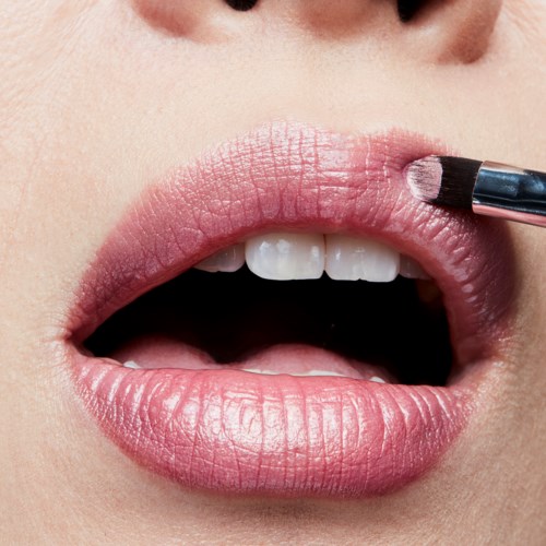 A person applies light pink lipstick to lips, with teeth showing slightly. Close-up focused on the mouth and lipstick brush.