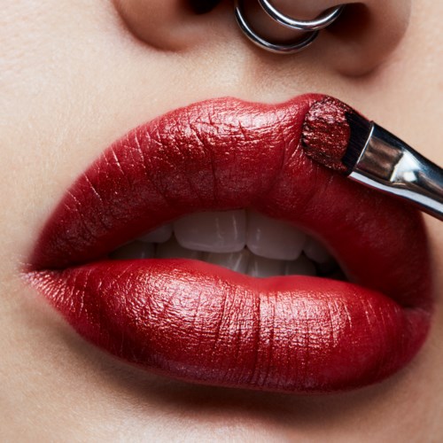 Close-up of lips being painted with red lipstick using a small brush, showing the lip piercing above.