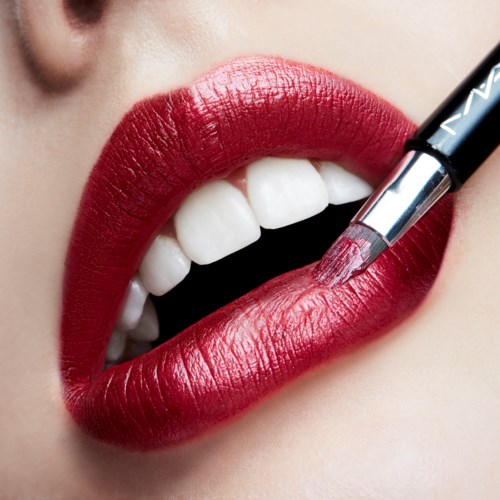 Close-up of lips being painted bright red with lipstick, showing teeth and a cosmetic brush.