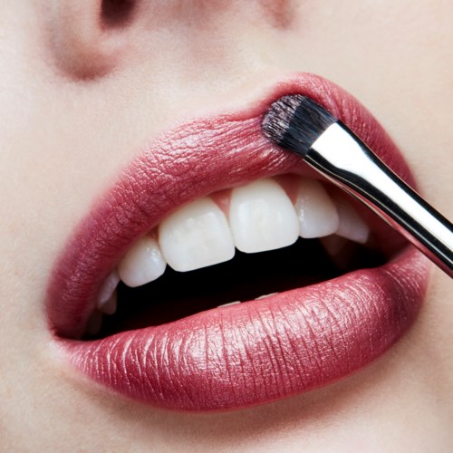 A person applying lipstick with a brush to their lips, showing close-up of mouth and teeth.