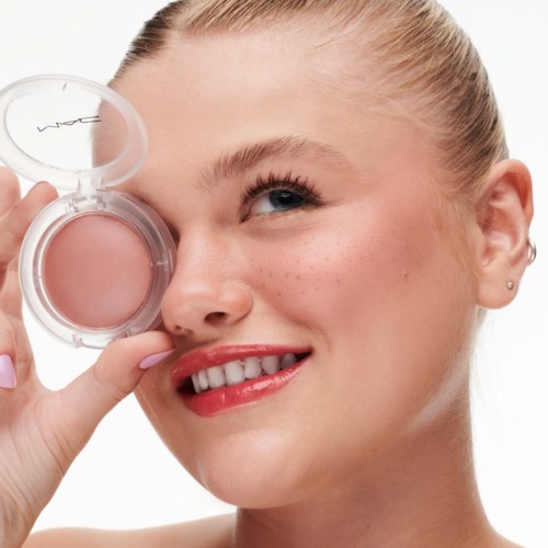 Eine Frau hält eine pinke Make-up-Box in der Nähe ihres Auges und lächelt. Sie hat blondes Haar und helle Haut mit Sommersprossen.