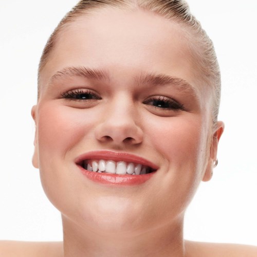 A young woman smiling with clear skin, blonde hair tied back, and bright eyes. She looks happy and confident.
