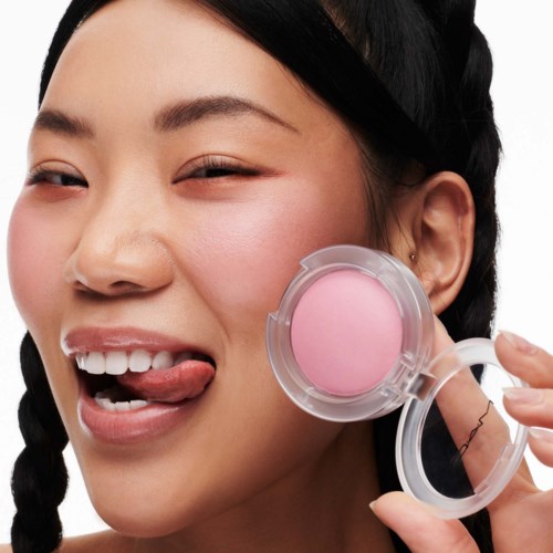 A woman with braided hair, smiling, licking her lips, holding a pink compact powder container near her face.