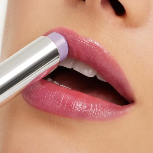 A person applies light purple lipstick to their lips, which are slightly parted showing white teeth.