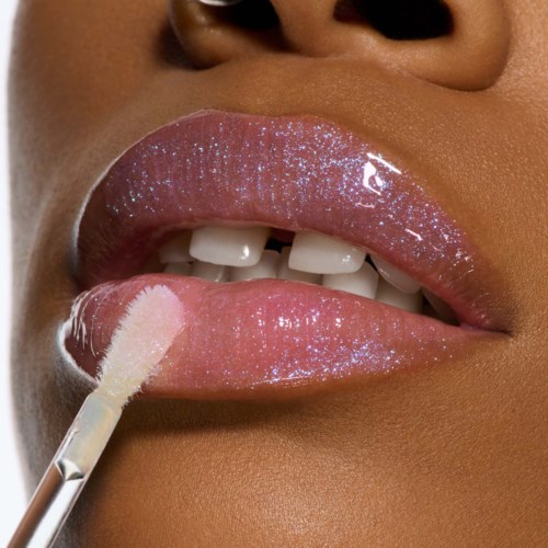 Close-up of lips with shiny pink gloss, a lip brush is applying gloss, showing teeth and a smooth skin tone.