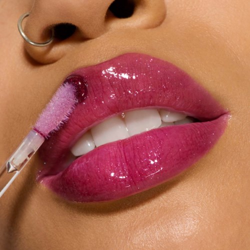 A person applies pink lip gloss with a brush, showing full lips and nose piercing.