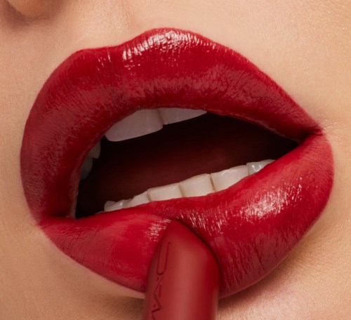 Close-up of a woman’s lips with bright red lipstick, slightly open showing teeth, and holding a lipstick stick.