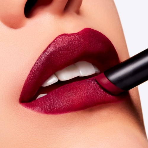 Close-up of lips with deep red lipstick, biting a matching lipstick tube.