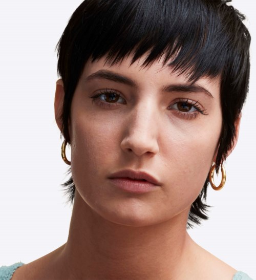 A woman with short, dark hair and earrings looks straight at the camera with a neutral expression.