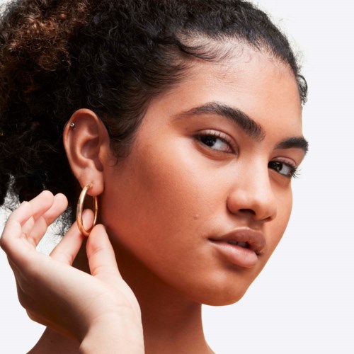 A woman with curly hair wearing gold hoop earrings, touching her earlobe with her hand.