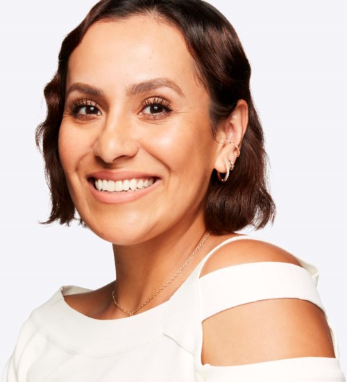A smiling woman with short brown hair, wearing earrings and a white top with shoulder cutouts.