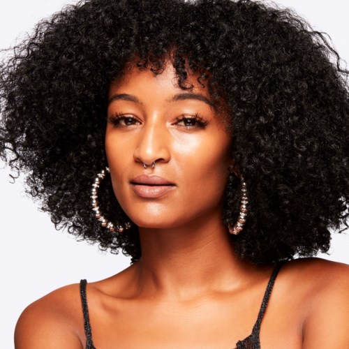 A woman with big, curly hair, wearing hoop earrings and a black strap top, looking confidently at the camera.