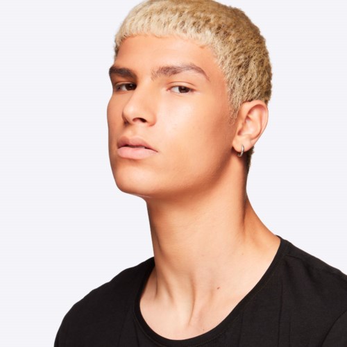 A young person with blonde hair and a piercing wears a black shirt, looking serious against a plain background.
