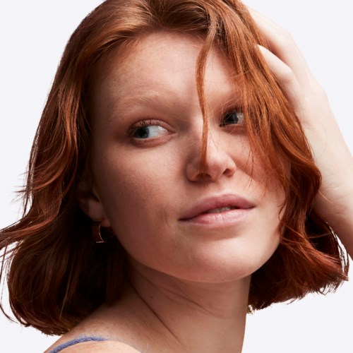 A woman with red hair is touching her head and looking to the side.
