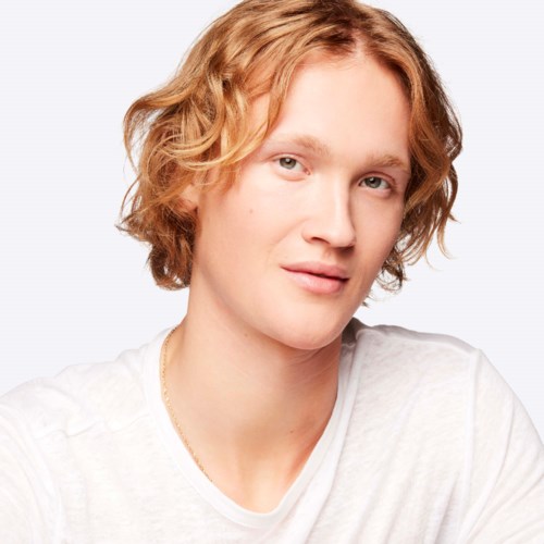 A young person with wavy, reddish-blonde hair, fair skin, and wearing a white shirt, smiling softly.