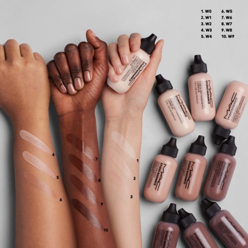 Two hands with different skin tones hold makeup foundation bottles, some swatched on arms with numbered labels. Several bottles lie nearby.