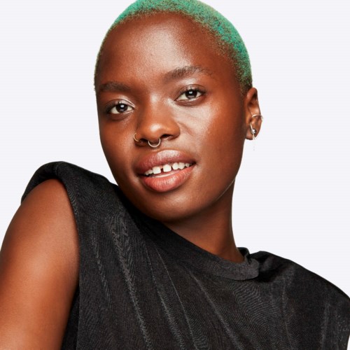 A young woman with short green hair, wearing a black sleeveless top, smiling gently, with a nose ring and earrings.